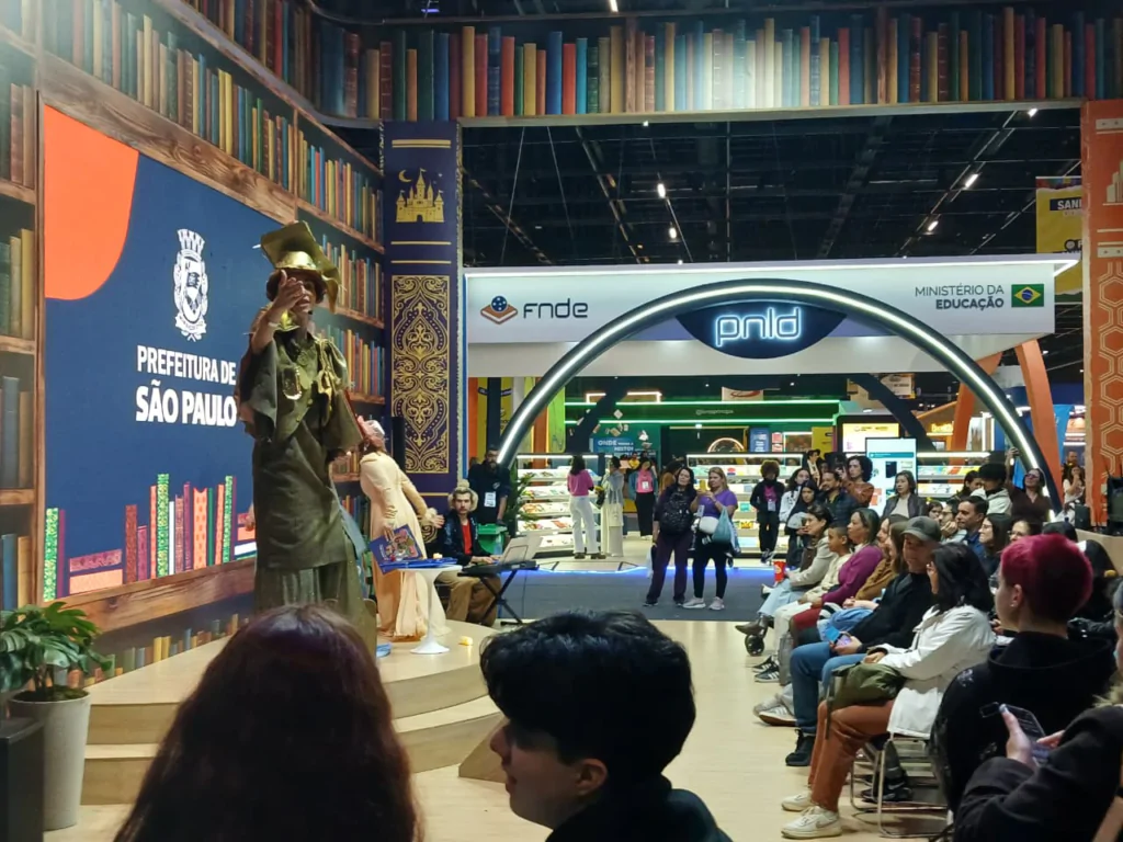Fotografia em plano geral de um evento cultural em ambiente fechado, mostrando uma apresentação teatral sobre um pequeno palco de madeira para uma plateia sentada. À esquerda, no palco sob um painel azul da "PREFEITURA DE SÃO PAULO" ladeado por estampas de prateleiras de livros, um ator fantasiado com trajes e chapéu em tom verde-oliva encena gesticulando para o público. Ao lado dele, outra artista veste trajes claros, enquanto um músico acomodado ao fundo acompanha a performance com um teclado. À direita e em primeiro plano, diversas pessoas assistem à apresentação sentadas em cadeiras dispostas em fileiras. Ao fundo, destaca-se uma estrutura em arco com os logotipos do "FNDE", "PNLD" e "MINISTÉRIO DA EDUCAÇÃO", com outros estandes e visitantes transitando pela feira.
