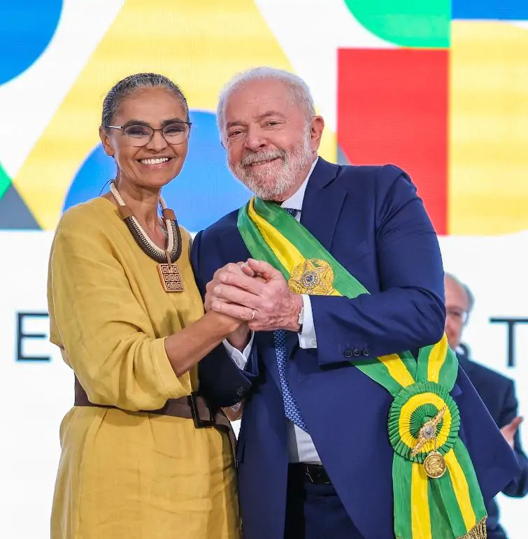 Mulher negra de óculos e roupa amarela com homem branco de terno azul e faixa presidencial