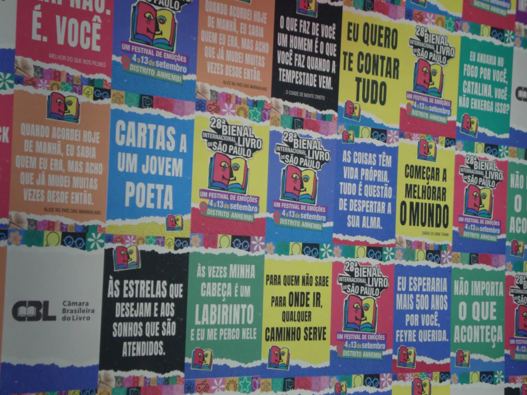 Fotografia em plano médio de uma parede inteiramente coberta por uma colagem de cartazes e painéis gráficos coloridos. Os cartazes exibem frases inspiradoras e literárias em letras garrafais brancas sobre fundos de cores variadas como verde, azul, vermelho e amarelo, intercalados com o logotipo oficial da "28ª Bienal Internacional do Livro de São Paulo".