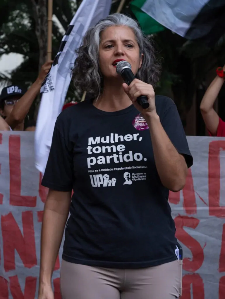 Mulher branca de cabelo branco e camiseta preta falando no microfone