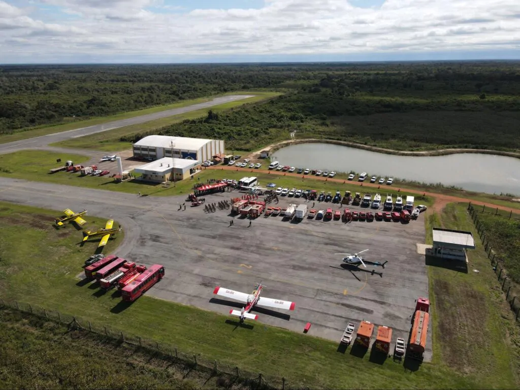 Foto aérea de um terrenos asfaltado em meio a uma vegetação com vários veículos estacionados.