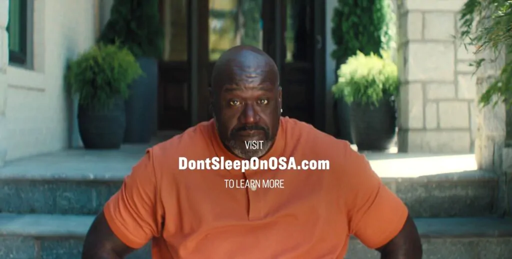 Foto do Shaquille O’Neal olhando fixo para a câmera com uma escada e vasos ao fundo.