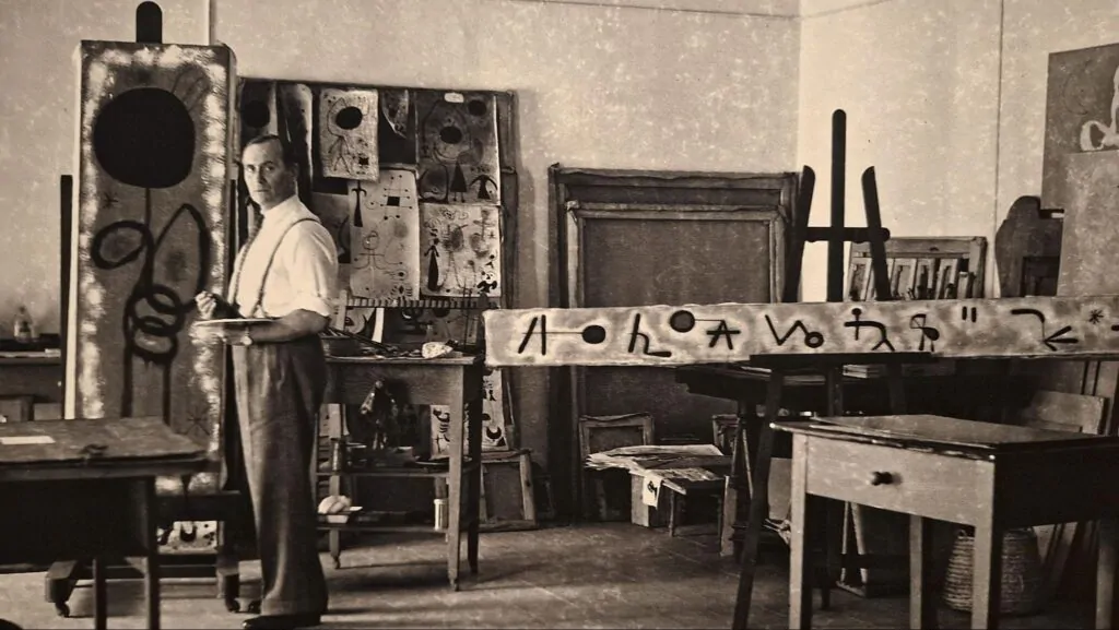 Foto de fotografia em preto e branco do artista surrealista Joan Miró em seu ateliê de arte cercado por telas e esboços.